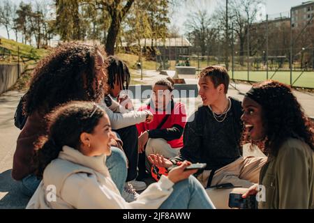 Multirassische männliche und weibliche Freunde sprechen, während sie zusammen im Park sitzen Stockfoto