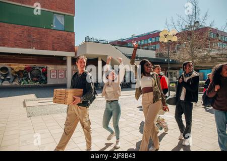 Glückliche multirassische Freunde winken die Hände, während sie an einem sonnigen Tag auf der Straße spazieren Stockfoto