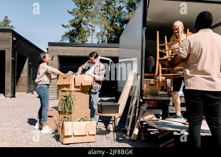 Professionelle Mover tragen Stühle vom LKW, während das Paar durch Kisten schaut Stockfoto