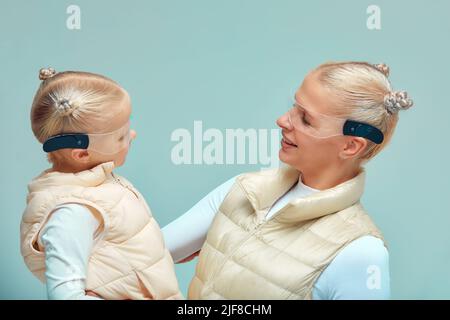 Virtuelle Technologie. Glücklich entzückt nettes Mädchen lächelnd und zu ihrer Mutter schauend, während Mutter in ihren Armen in der digitalen virtuellen Brille auf hält Stockfoto