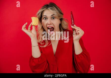 Frau in rotem Kleid auf rotem Hintergrund schreit laut und hält Chilischoten und Pommes in den Händen. Würziges Essen. Stockfoto
