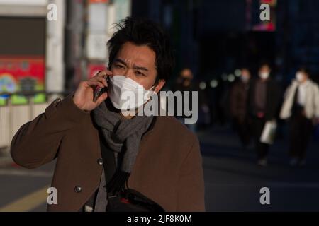 Ein Japaner, der eine OP-Maske trägt, spricht auf seinem Handy, während er in Shinjuku, Tokio, Japan, auf der Straße geht. Stockfoto