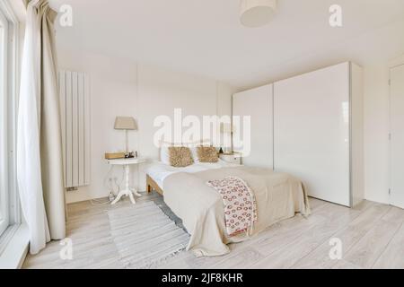 Komfortables Bett mit gepunkteter Federdecke in minimalistischem Stil in der Nähe des Kleiderschrankes im modernen Apartment Stockfoto