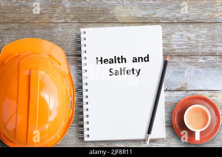 Gesundheit und Sicherheit mit orangefarbenem Helm, Bleistift, Kaffeetasse und Notizbüchern auf Holztisch am Büroarbeitsplatz. Stockfoto