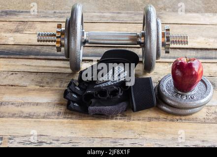 Roter Apfel und Hanteln, zusätzliche Gewichte und schwarze Handschuhe auf dem Holzboden Stockfoto