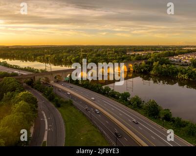 Luftaufnahme der Eisenbahnbrücke über den Raritan-Fluss in New Brunswick, New Jersey, während der Sommeruntergang die Bögen der Brücke beleuchtet Stockfoto