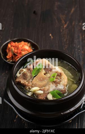 Gori Gomtang oder koreanische Rinderoxtail-Eintaufsuppe, serviert in der schwarzen koreanischen Schüssel mit Kimchi und geschnittenen grünen Zwiebeln auf dem schwarzen Holztisch Stockfoto