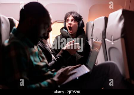 Eine multiethnische Gruppe von Menschen, die in der Economy Class mit Laptop, Smartphone und Kopfhörern während des Sonnenuntergangs ins Ausland fliegen. Passagiere, die mit einer internationalen kommerziellen Fluggesellschaft reisen. Stockfoto