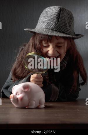 Der niedliche kleine Detektiv mit Lupe schaut auf das Sparschwein. Ein kleines Mädchen in Hut und Mantel spielt Detektiv. Stockfoto