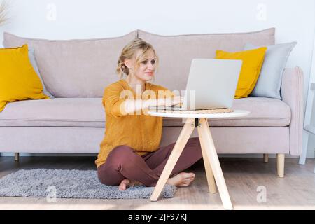 Seitenansicht. Schöne Frau überprüft E-Mails online, nutzt einen modernen Laptop, arbeitet aus der Ferne, nutzt WLAN. Nette lächelnde Frau, die auf dem Boden neben dem Sofa zu Hause auf weißem Hintergrund sitzt. Speicherplatz kopieren Stockfoto