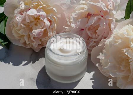 Geöffnetes weißes Glas mit leichter Kräutercreme für Frauen auf einem Marmortisch. Schönes Bouquet von blühenden Pfingstrosen. Pflege für sauberes und weiches Gesicht, Hände, le Stockfoto