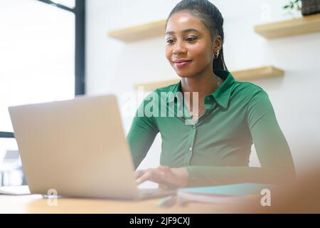 Bezaubernde junge afroamerikanische Geschäftsfrau, die ihren Laptop für die Büroarbeit trägt, weibliche Angestellte überprüft die Post und führt Geschäftskorrespondenz mit Kunden Stockfoto