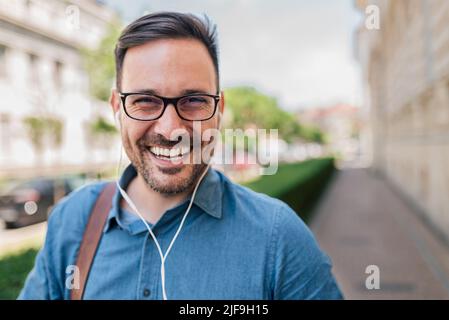 Lächelnder junger Geschäftsmann mit Kopfhörern. Porträt eines männlichen professionellen Pendlers, der Musik hört. Er trägt in der Stadt Brillen und Formalien. Stockfoto