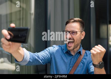 Glücklicher Geschäftsmann, der Selfie auf dem Smartphone macht. Der männliche Vorstandskleider klemmt beim Schreien die Faust. Er steht gegen das Bürogebäude in der Stadt. Stockfoto