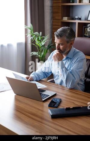 Porträt eines erwachsenen grauhaarigen bärtigen Geschäftsmannes, der im Heimbüro mit einem Laptop arbeitet. SMS senden, Schreibarbeiten erledigen, monatliche Ausgaben berechnen Stockfoto