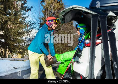 Mutter helfen Sohn auf Snowboard Stiefel sitzen im Kofferraum zu setzen Stockfoto