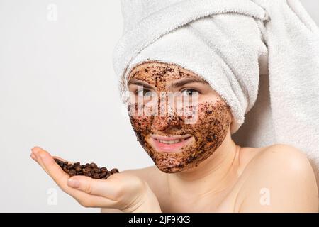 Eine Frau mit einer Kaffeemaske oder einem Peeling im Gesicht hält eine Handvoll Kaffeebohnen in der Hand Stockfoto