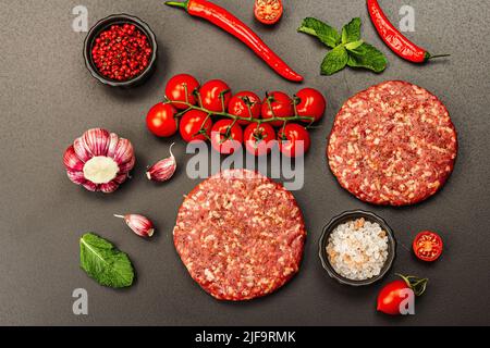 Rohe Steak-Koteletts aus Hackfleisch auf schwarzem Stein-Hintergrund. Frisches Gemüse, Minze und Gewürze. Traditionelle Zutat für einen Burger, flaches Lay, Stockfoto