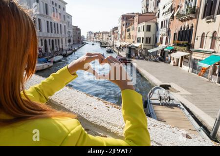 Teilansicht einer rothaarige Frau, die ein Herzzeichen über dem verschwommenen Canal Grande in Venedig zeigt Stockfoto