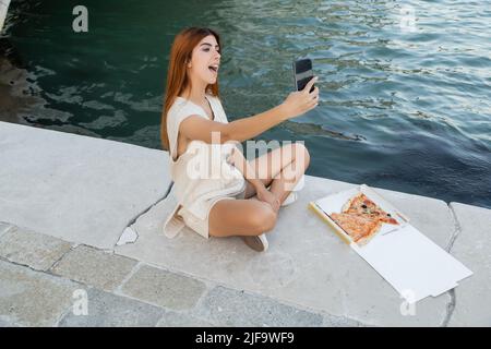 Aufgeregt Frau, die Selfie nimmt, während sie in der Nähe einer Kartonschachtel mit leckerer Pizza auf dem Bürgersteig in Venedig sitzt Stockfoto