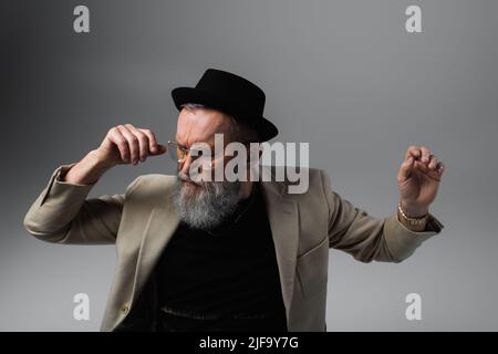 Bärtiger älterer Mann mit Derby-Hut und Brillen, die auf Grau gestikulierten Stockfoto