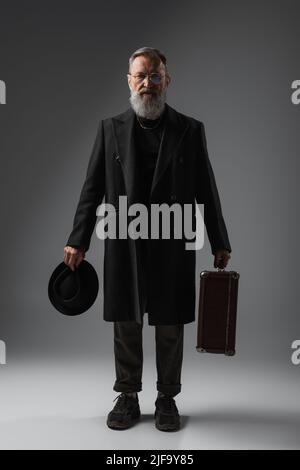 In voller Länge von eleganten Senior Mann in Mantel hält Derby Hut und Koffer auf grau Stockfoto