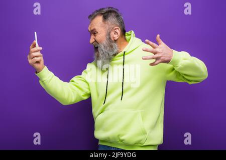 Gestresster älterer Mann im grünen Hoodie, der Smartphone auf lila anschaut Stockfoto