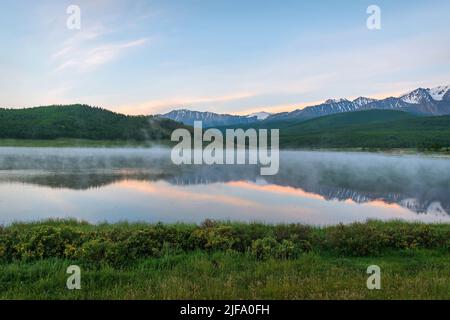 Erstaunlich zarte Morgendämmerung über den Bergen mit Schnee und Wald bedeckt, Nebel über dem See und schöne Reflexionen im Wasser gegen den blauen Himmel a Stockfoto