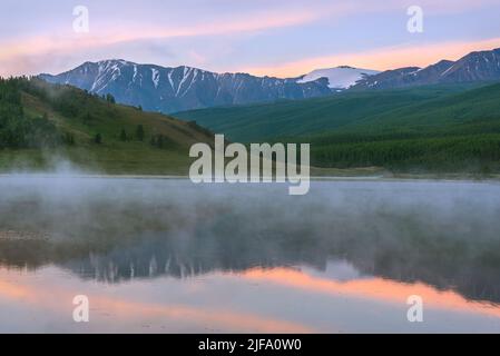 Erstaunlich zarte Morgendämmerung über den Bergen mit Schnee und Wald bedeckt, Nebel über dem See und schöne Reflexionen im Wasser gegen den blauen Himmel a Stockfoto