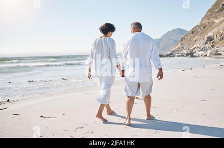 Rückansicht eines Seniorenpaares mit gemischtem Rennen, das einen romantischen Spaziergang am Strand Unternehmen und sich an einem sonnigen Sommertag im Freien die Hände halten kann Stockfoto