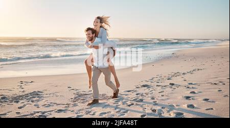 Glückliches Paar, das am Strand spielt. Unbeschwertes junges Paar, das gemeinsam Spaß im Urlaub hat. Ehemann, der seiner Frau eine Huckepack-Fahrt gab. Verheiratetes Paar Stockfoto