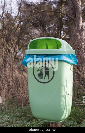 Leeren Sie den hellgrünen Mülleimer oder Mülleimer mit einem blauen Müllbeutel und einem Piktogramm im Winter in einem Wald oder Park mit Bäumen im Hintergrund Stockfoto
