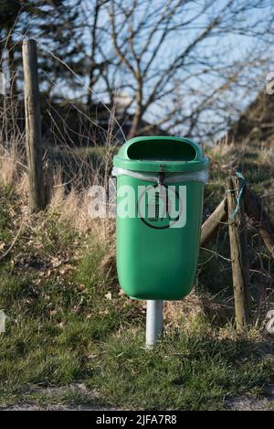 Leerer grüner Mülleimer mit Müllbeutel und einem Piktogramm, das neben einem Zaun in einer ländlichen Gegend steht - aufrechtes Bild, aufgenommen an einem sonnigen Nachmittag im Winter Stockfoto