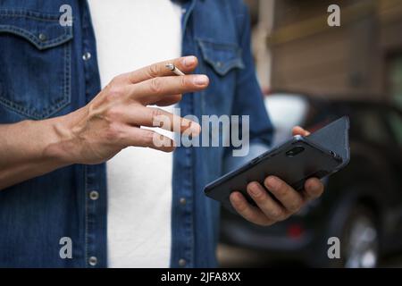 Ein Mann im mittleren Alter, der Zigarette raucht, überprüft SMS oder soziale Medien mit seinem Smartphone, das Jeans-Hemd und weißes T-Shirt trägt und draußen im Stadthintergrund steht. Kein Gesicht sichtbar. Stockfoto