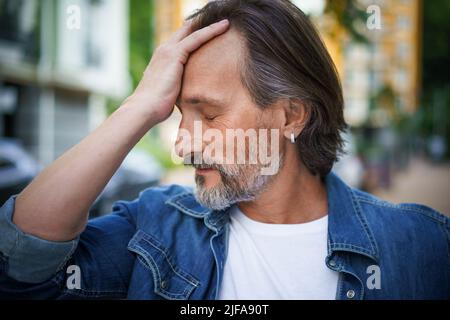 Ein gutaussehender Mann übergeht die Krise des mittleren Alters und berührt seine Stirn, die emotional erschöpft aussieht. Kopfschmerzen, Stress reifer schöner Mann im Freien europäischen alten Stadtstraßen stehen. Stockfoto