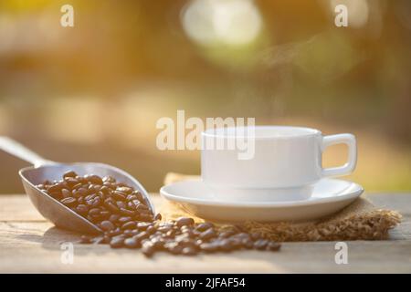 Nahaufnahme einer Tasse Kaffee und Kaffeebohnen auf dem Holztisch im Freien bei morgendlichem Sonnenlicht und Bokeh-Hintergrund Stockfoto