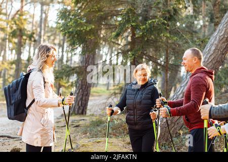 Lächelnde Sporttrainerin mit einer Gruppe von Leuten, die sich unterhalten und die Haltung des Nordic Walking mit Stöcken im Wald erklären Stockfoto