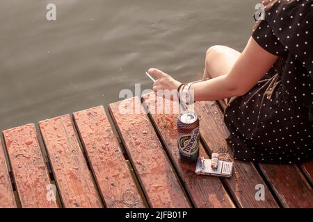 Bild von den Händen einer jungen Frau, die mit einer Bierkachtel Alkohol trinkt und mit einer Zigarette Tabak raucht, an einem Fluss in Belgrad, Serbien Stockfoto