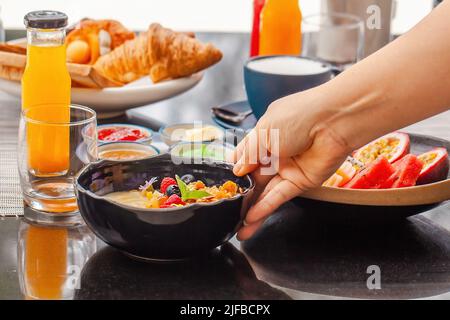 Gesundes veganes Essen im Café. Frau Hand legt einen Teller mit Smoothie Schüssel auf den Tisch. Morgens Bio-Lebensmittel, frisches Obst, Säfte, laktosefreier Latte Stockfoto