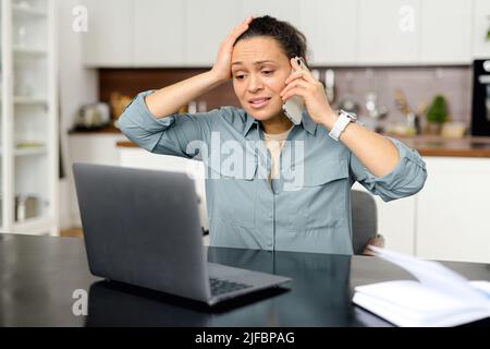 Verärgert und frustriert Frau mit Laptop für Remote-Arbeit von zu Hause aus, nervös über Fehler, stressigen Job, gemischt Rennen Frau sprechen auf dem Smartphone, hat Problem mit der Aufgabe Stockfoto