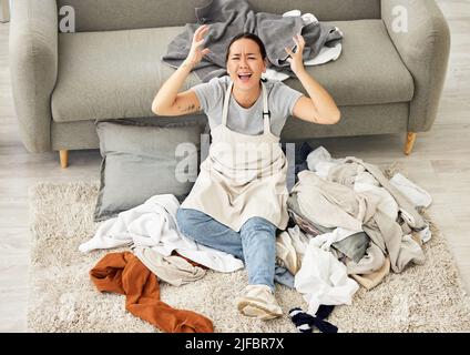 Eine Frau mit gemischter Rasse, die in einem Haufen schmutziger Wäsche sitzt und im Wohnzimmer einen Nervenzusammenbruch hat. Eine junge asiatische Frau fühlt sich überwältigt und Stockfoto