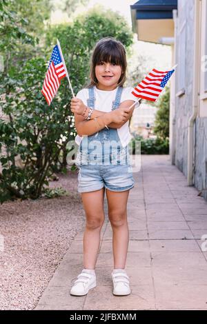 Kleines schwarzhaariges Mädchen mit amerikanischen Flaggen im Garten ihres Hauses. Konzept der Feier, Unabhängigkeitstag, 4.. Juli, Patriotismus, Feiertag und Stockfoto