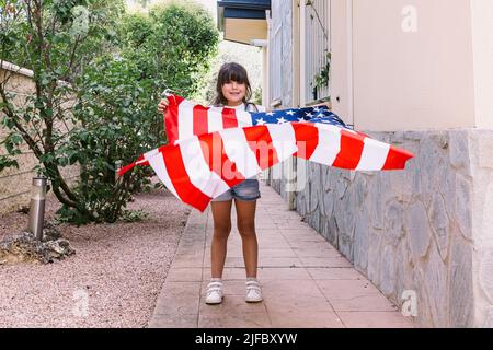 Das schwarzhaarige Mädchen hält und schwenkt eine Flagge der Vereinigten Staaten im Garten ihres Hauses. Konzept der Feier, Unabhängigkeitstag, Vereinigte Staaten von Amer Stockfoto