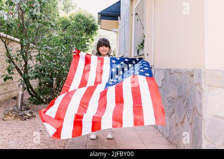 Das schwarzhaarige Mädchen hält und schwenkt eine Flagge der Vereinigten Staaten im Garten ihres Hauses. Konzept der Feier, Unabhängigkeitstag, Vereinigte Staaten von Amer Stockfoto