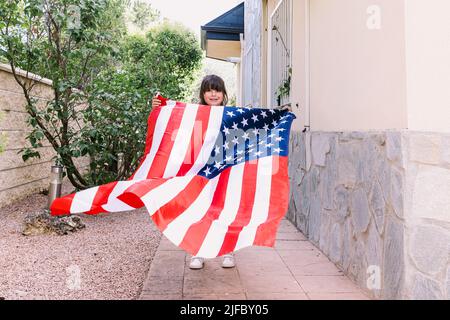 Das schwarzhaarige Mädchen hält und schwenkt eine Flagge der Vereinigten Staaten im Garten ihres Hauses. Konzept der Feier, Unabhängigkeitstag, Vereinigte Staaten von Amer Stockfoto