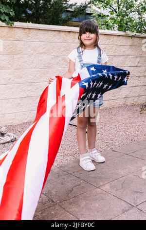 Das schwarzhaarige Mädchen hält und schwenkt eine Flagge der Vereinigten Staaten im Garten ihres Hauses. Konzept der Feier, Unabhängigkeitstag, Vereinigte Staaten von Amer Stockfoto