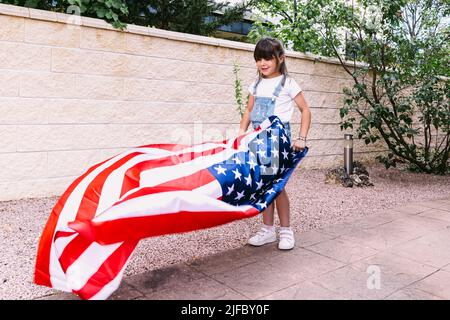 Das schwarzhaarige Mädchen hält und schwenkt eine Flagge der Vereinigten Staaten im Garten ihres Hauses. Konzept der Feier, Unabhängigkeitstag, Vereinigte Staaten von Amer Stockfoto