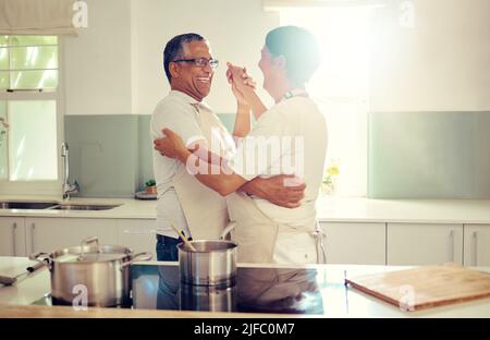 Ein liebevolles Senioren-Mischpärchen trägt Schürzen und hat Spaß beim Stehen und Tanzen in der Küche zu Hause. Romantische reife Mann und Frau lächeln und Stockfoto