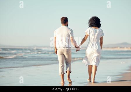 Rückansicht eines jungen Paares mit gemischter Rasse, das sich beim gemeinsamen Spaziergang am Strand die Hände hielt. Hispanische Paare reisen und genießen Urlaub und sein Stockfoto