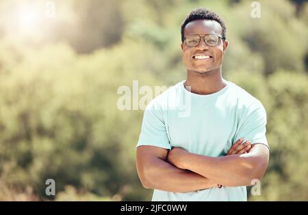 Ein hübscher junger afroamerikanischer Mann, der eine Brille trägt und draußen mit gefalteten Armen steht. Glücklicher und selbstbewusster schwarzer Mann lächelt während Stockfoto
